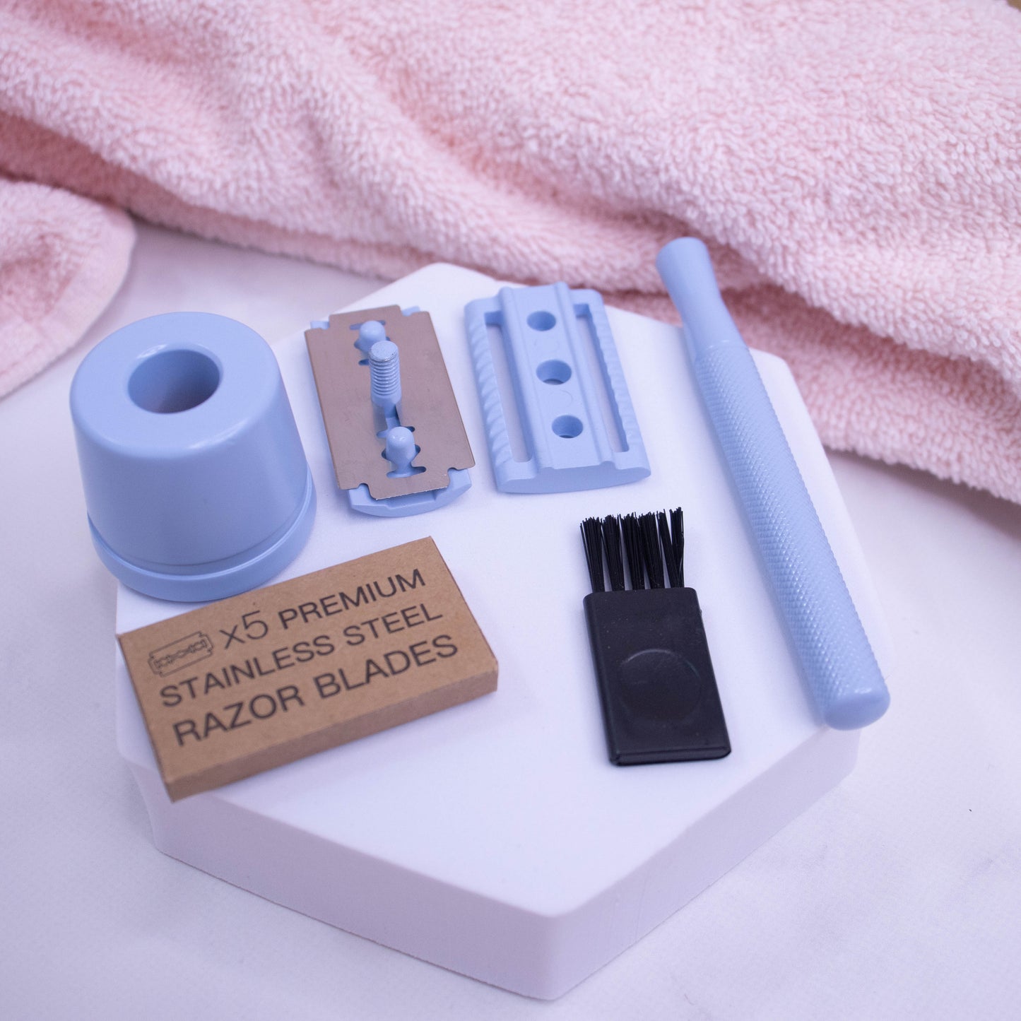 A pink towel lies by a white foam stand with a dismantled pale blue safety razor. It demonstates how the handle twists off the head of the razor which splits into two with a silver blade resting on one half of the head. A pack of five razor blades in cardboard box and small black cleaning brush also rest on the white surface.