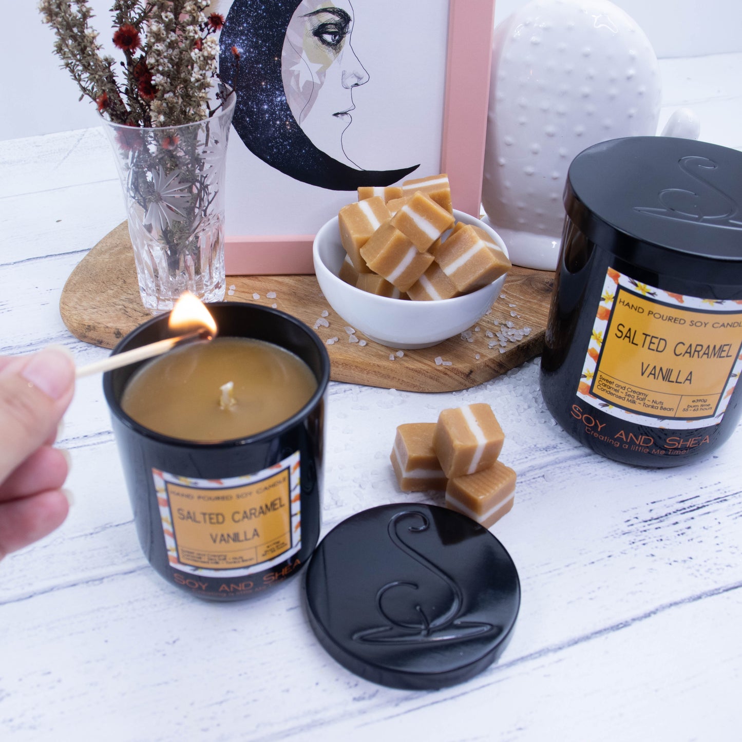Two candles in black glass jars, with metal lids and decorative labels stand on a white wooded surface. A larger looking candle sits to the right of the image with its lid on, and smaller one sits to the left with its lid removed and placed on the side revealing a caramel coloured wax. A hand reaches in to light the candle with a match. In the background is a small white bowl of caramel pieces with sea salt scattered around, sitting next to a picture frame and vase of dried flowers.