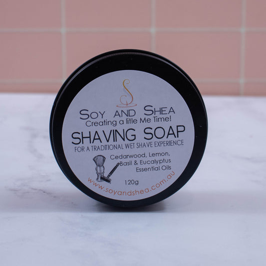 A round black tin stands on its side on a marble bench with peach tile background. The lid of the tin has a label showing the details of the Shaving soap