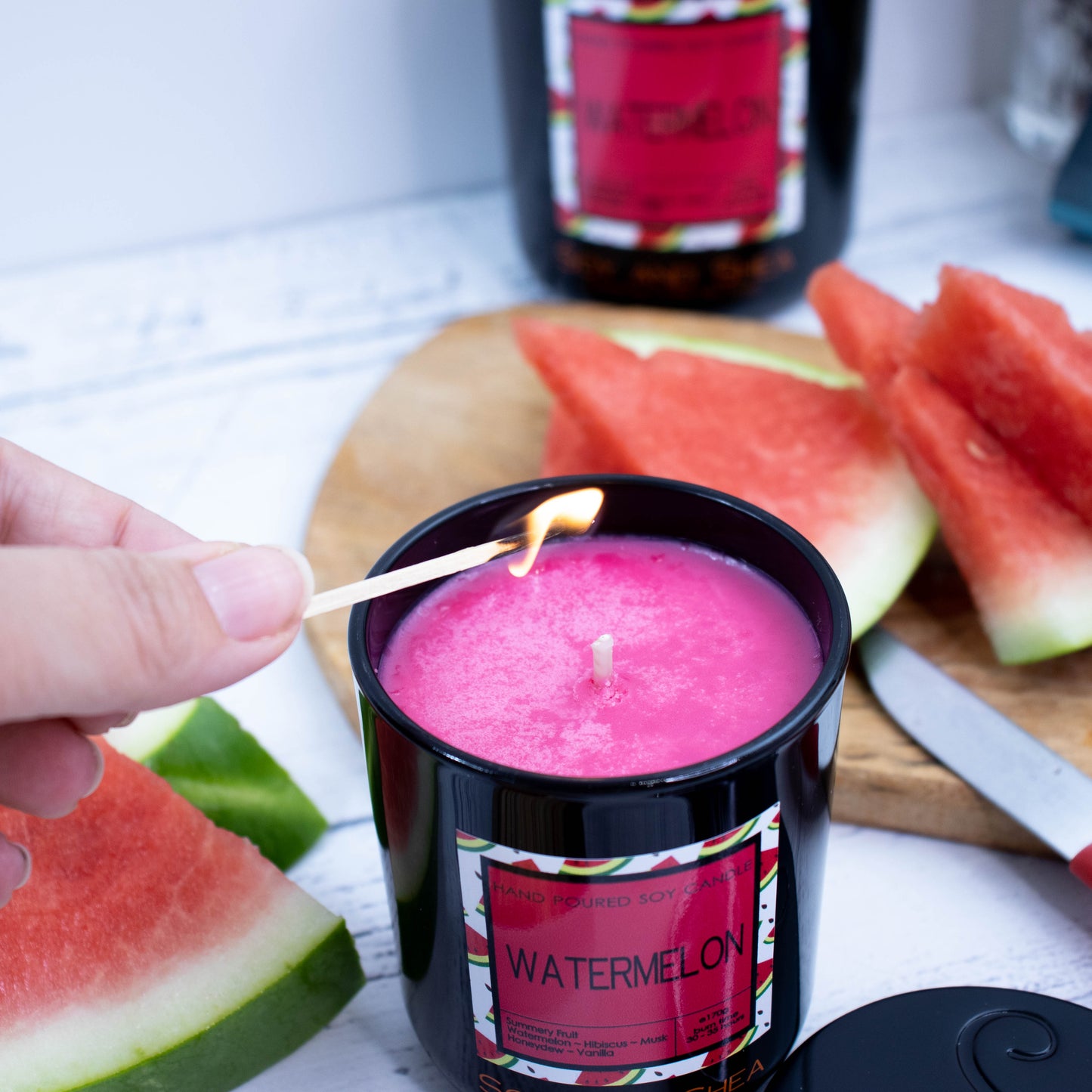 A black glass jar candle sits in the foreground with its metal lid sitting to the side revealing a deep pink wax. A Hand reaches in to light the candle with a match. Surrounding the candle are slices of watermelon and in the blurred background sits another large candle