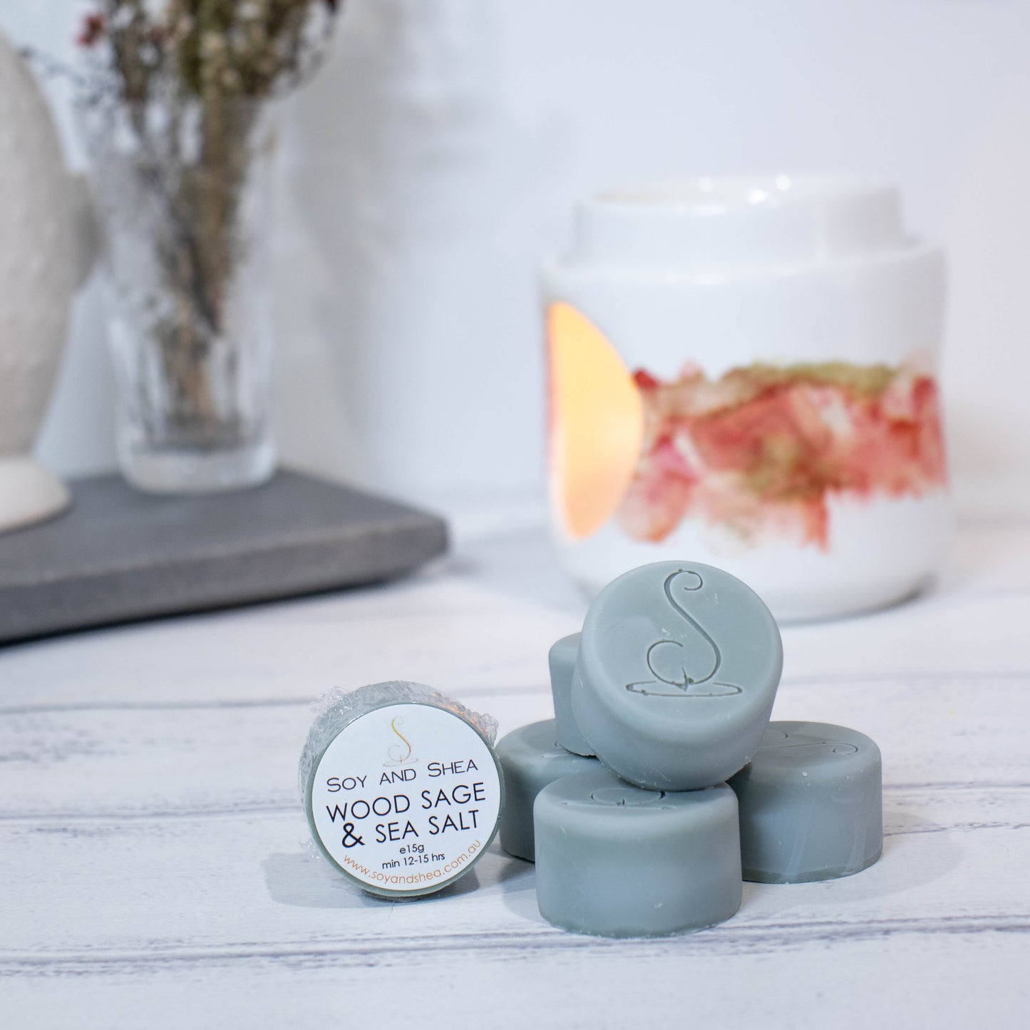 Four cylindrical shaped Melt Pucks in moss green are stacked up on each other in pyramid form with the soy and shea logomark stamped on the tops. To the left is a fourth melt puck that rests on its side with a label identifying it as Wood Sage and Sea Salt. These sit on a wooden surface with a lit fragrance warmer in the backgroung along with a vase of flowers.