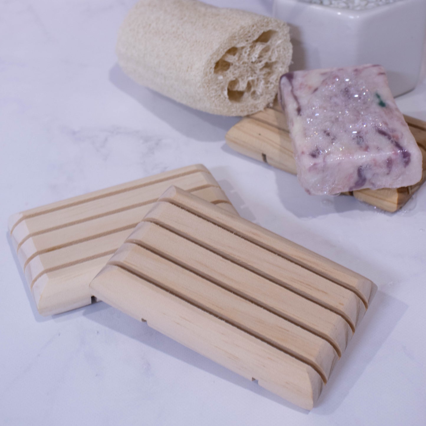 Two wooden soap dishes rest against each other on a marble surface. Behind sits a third wooden soap dish with a soap bar with bubbles next to a natural loofah.