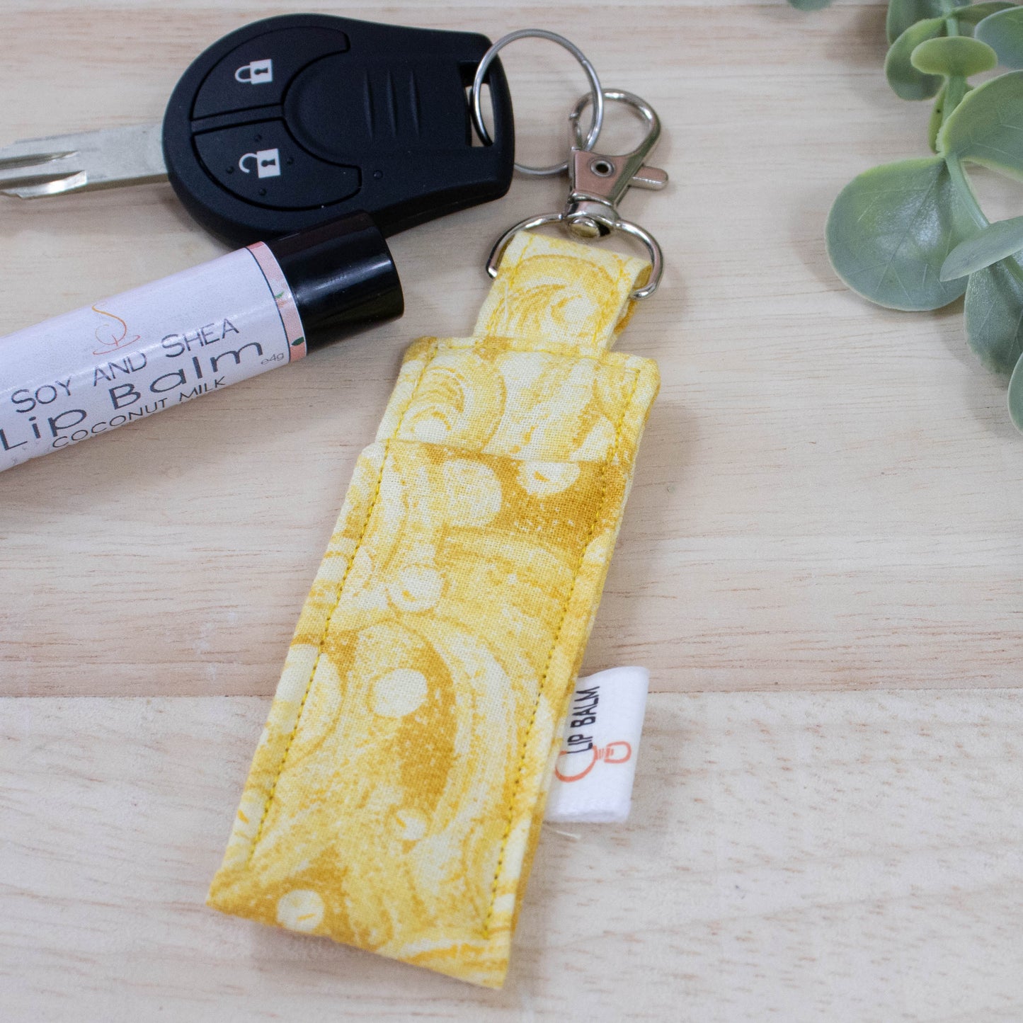 A clip balm holder lies on a wooden surface attached to a car key with a "lobster claw" clip. To the side is a tube of lip balm and some greenery. The Clip Balm has a wave like pattern in shades of yellow.