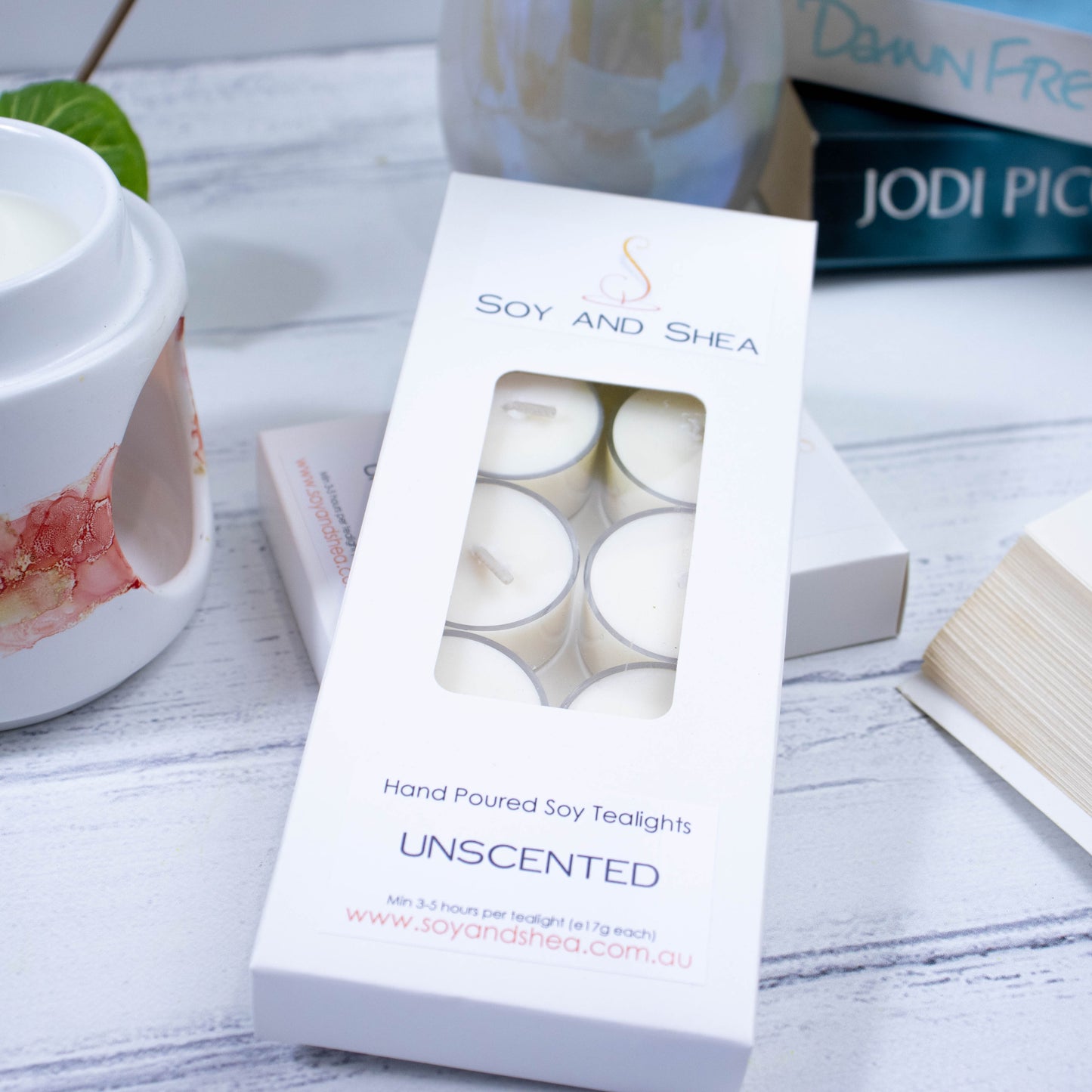 A long rectagular box of Unscented Soy Tea Lights by Soy and Shea displayed alongside a tea light holder and a book, all placed on a white wooden surface. The box is white with window cutout showing polycarbonate tealight cups filled with white wax.
