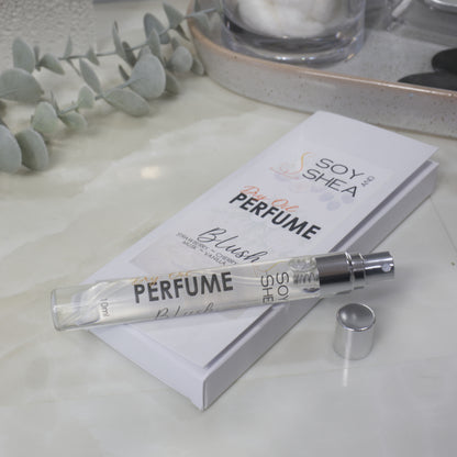 A tall, narrow clear perfume bottle lays on top of its box packaging identifying it as Dry Oil Perfume, Blush.  The bottle is labelled as 10ml and features a silver spray nozzle with the silver cap resting on the side.  In background is some greenery.