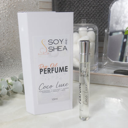 A tall, narrow, clear perfume bottle with a silver cap stands upright on a bathroom counter top next to it's packaging.   The packaging is a white box labelled as Dry Oil Perfume  Coco Luxe