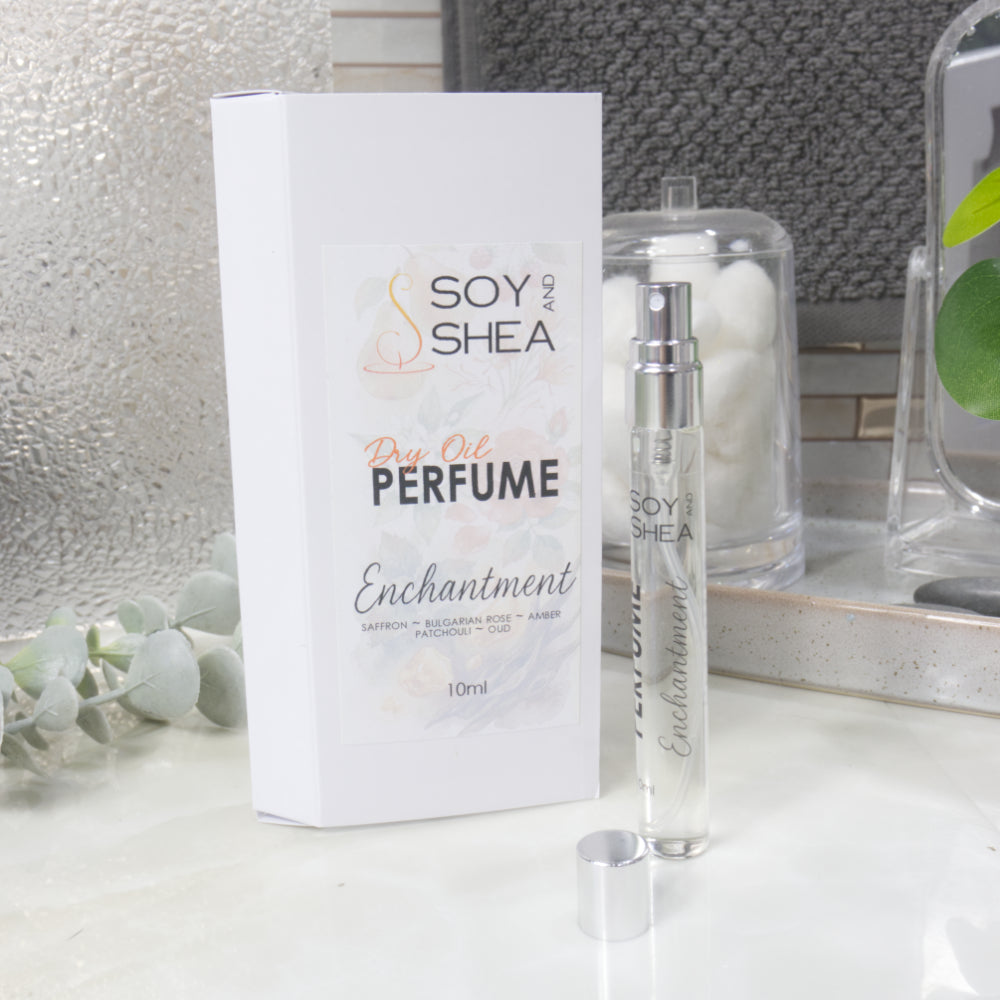 A tall, narrow, clear perfume bottle with a silver atomonise stands upright on a bathroom counter top next to it's packaging and the silver cap.   The packaging is a white box labelled as Dry Oil Perfume Enchantment