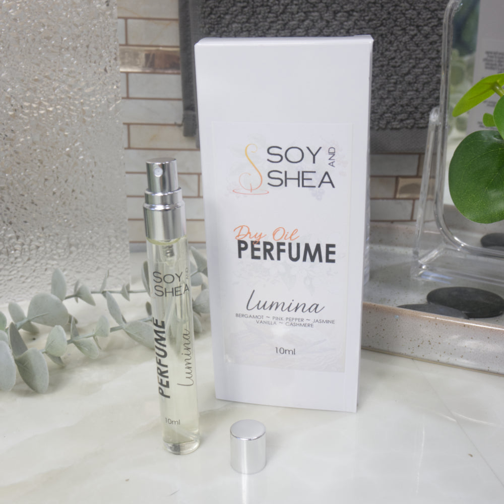 A tall, narrow, clear perfume bottle with a silver atomiser stands upright on a bathroom counter top next to it's packaging and a silver cap.   The packaging is a white box labelled as Dry Oil Perfume Lumina