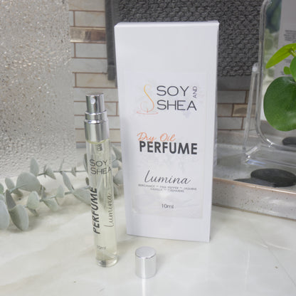 A tall, narrow, clear perfume bottle with a silver atomiser stands upright on a bathroom counter top next to it's packaging and a silver cap.   The packaging is a white box labelled as Dry Oil Perfume Lumina