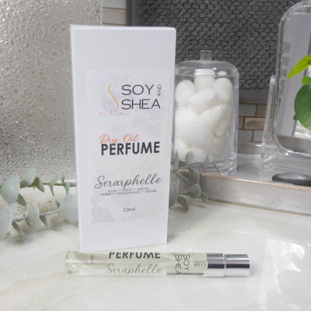 A long, narrow, clear bottle of Dry Oil Perfume with a silver cap lays on a bathroom countertop in front of its packaging box.  Behind is other bathroom accessories