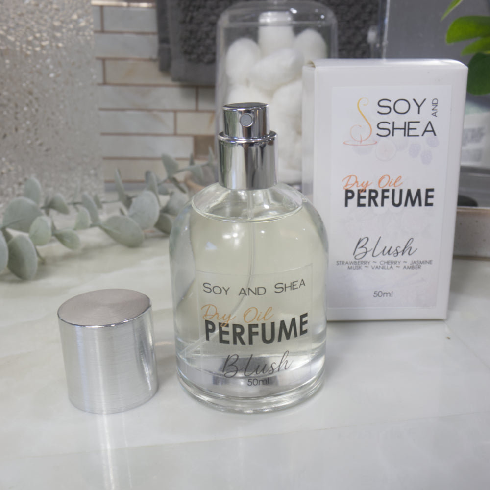 A squat, clear round bottle sits on a bathroom counter top with a clear liquid inside.  The bottle has a silver cap that has been removed and placed to the side revealing a silver atomiser.  The white packaging box sits behind the bottle labelled as Dry Oil Perfume Blush.