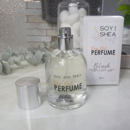 A squat, clear round bottle sits on a bathroom counter top with a clear liquid inside.  The bottle has a silver cap that has been removed and placed to the side revealing a silver atomiser.  The white packaging box sits behind the bottle labelled as Dry Oil Perfume Blush.