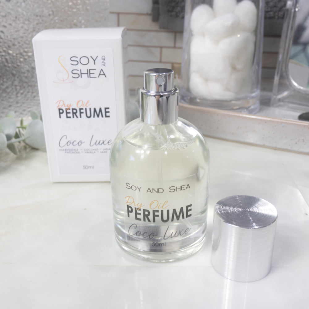 A squat, clear round bottle sits on a bathroom counter top with a clear liquid inside.  The bottle has a silver cap that has been removed and placed to the side revealing a silver atomiser.  The white packaging box sits behind the bottle labelled as Dry Oil Perfume Coco Luxe.
