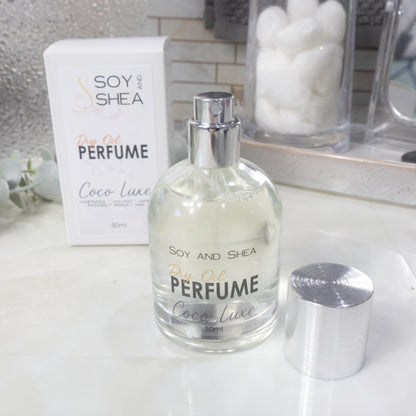 A squat, clear round bottle sits on a bathroom counter top with a clear liquid inside.  The bottle has a silver cap that has been removed and placed to the side revealing a silver atomiser.  The white packaging box sits behind the bottle labelled as Dry Oil Perfume Coco Luxe.