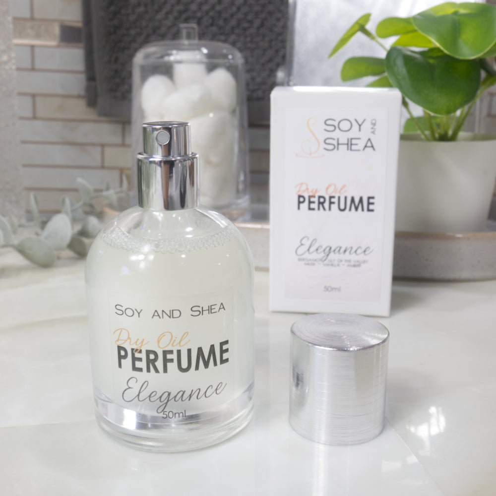 A squat, clear round bottle sits on a bathroom counter top with a cloudy clear liquid inside.  The bottle has a silver cap that has been removed and placed to the side revealing a silver atomiser.  The white packaging box sits behind the bottle labelled as Dry Oil Perfume Elegance.