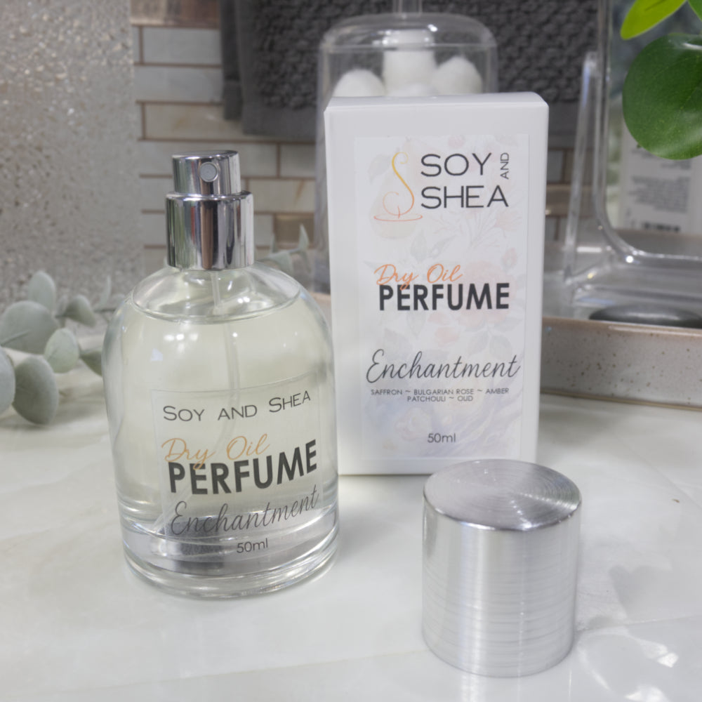 A squat, clear round bottle sits on a bathroom counter top with a clear liquid inside.  The bottle has a silver cap that has been removed and placed to the side revealing a silver atomiser.  The white packaging box sits behind the bottle labelled as Dry Oil Perfume Enchantment.