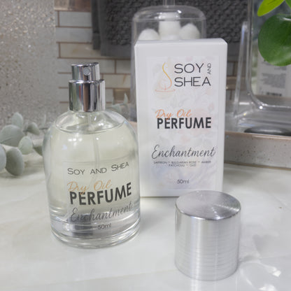 A squat, clear round bottle sits on a bathroom counter top with a clear liquid inside.  The bottle has a silver cap that has been removed and placed to the side revealing a silver atomiser.  The white packaging box sits behind the bottle labelled as Dry Oil Perfume Enchantment.