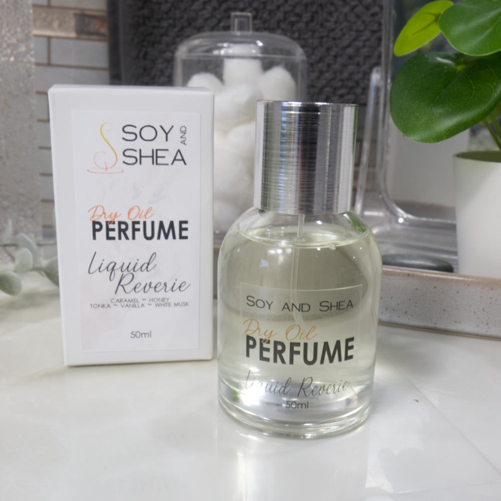 A squat, clear round bottle sits on a bathroom counter top with a clear liquid inside.  The bottle has a silver cap that has been removed and placed to the side revealing a silver atomiser.  The white packaging box sits behind the bottle labelled as Dry Oil Perfume Liquid Reverie.