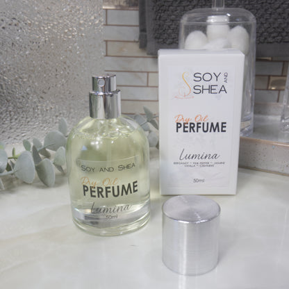A squat, clear round bottle sits on a bathroom counter top with a clear liquid inside.  The bottle has a silver cap that has been removed and placed to the side revealing a silver atomiser.  The white packaging box sits behind the bottle labelled as Dry Oil Perfume Lumina.