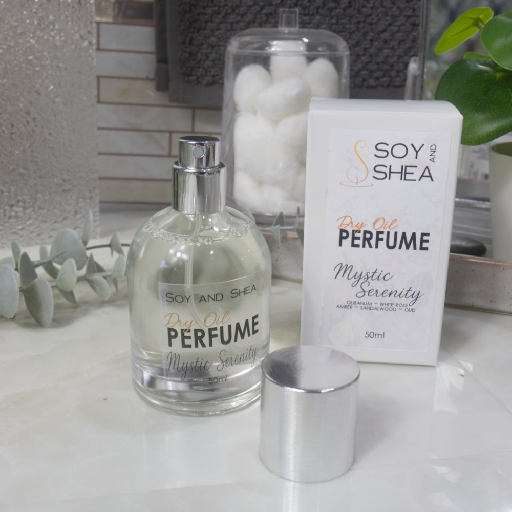 A squat, clear round bottle sits on a bathroom counter top with a clear liquid inside.  The bottle has a silver cap that has been removed and placed to the side revealing a silver atomiser.  The white packaging box sits behind the bottle labelled as Dry Oil Perfume Mystic Serenity.