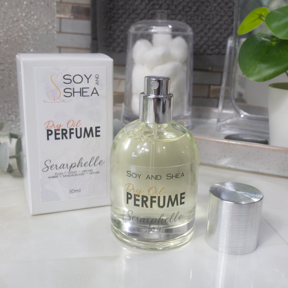 A squat, clear round bottle sits on a bathroom counter top with a clear liquid inside.  The bottle has a silver cap that has been removed and placed to the side revealing a silver atomiser.  The white packaging box sits behind the bottle labelled as Dry Oil Perfume Seraphelle.