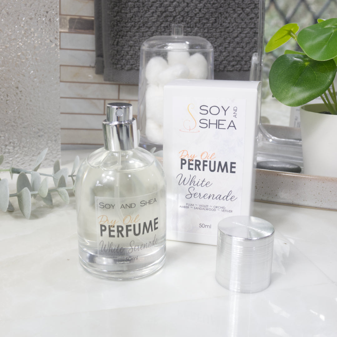 A squat, clear round bottle sits on a bathroom counter top with a clear liquid inside.  The bottle has a silver cap that has been removed and placed to the side revealing a silver atomiser.  The white packaging box sits behind the bottle labelled as Dry Oil Perfume White Serenade.