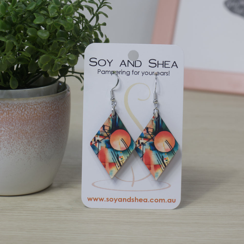 Diamond shaped earrings with an colourful blue/orange abstract design on a Soy and Shea branded card with a plant in the background