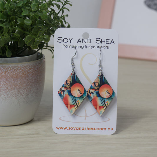 Diamond shaped earrings with an colourful blue/orange abstract design on a Soy and Shea branded card with a plant in the background