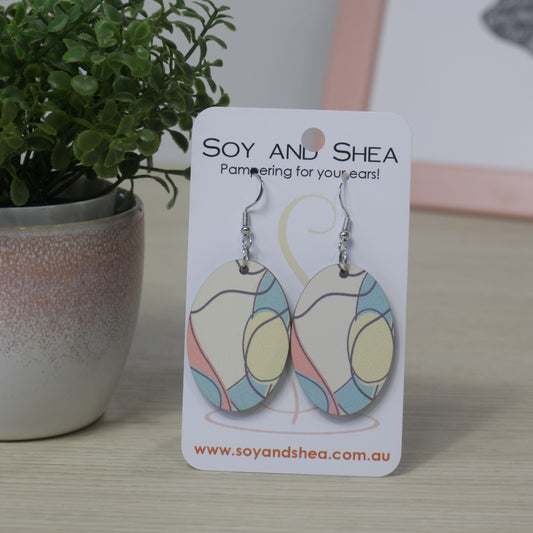 Oval shaped earrings with an abstract design of black squiggly lines with some areas colour light pink, blue or yellow on a Soy and Shea card with a plant in the background