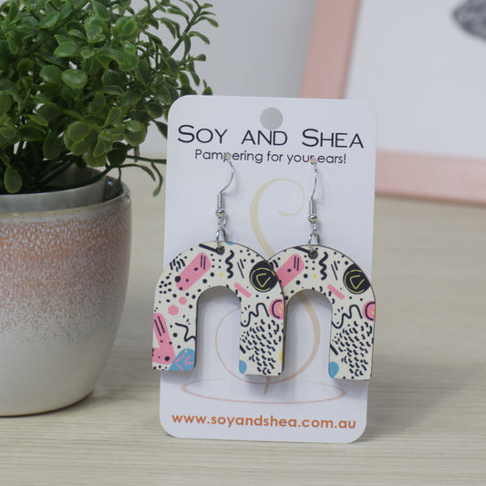 Pair of rainbow arched earrings on a 'Soy and Shea' branded card with a plant in the background.  The earrings feature an abstract pattern of black squiggles along with blue and pink spots.  