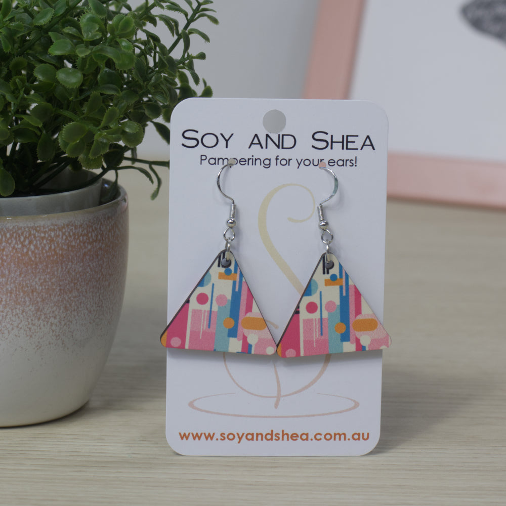 Colorful triangle-shaped earrings on a Soy and Shea branded card with a plant in the background.