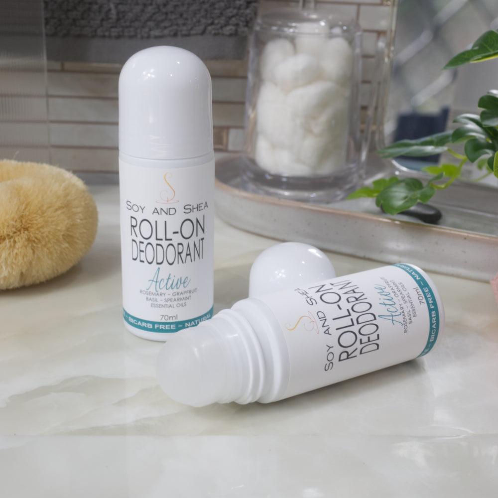 Two bottles of Roll-On Deodorant are displayed on a bathroom counter top with a jar of cotton balls, body brush and greenery.  One bottle stands upright with a domed cap, while another lays flay with the cap removed showing a ball roller in the top