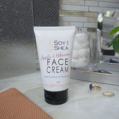 A white squeeze tube of Soy and Shea Face Cream with a black cap, placed on a marble countertop with a peach towel, rings, and a glass jar of cotton pads in a bathroom setting.