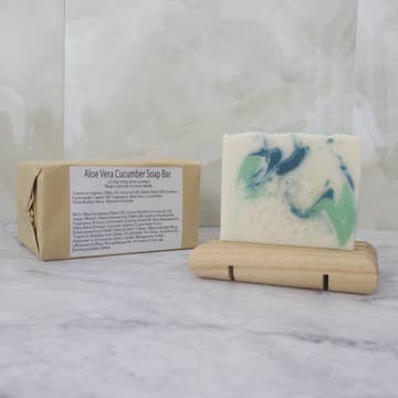 A white soap bar with green drops sits on a wooden stand with a brown paper package behind labeled 'Aloe Vera Cucumber Soap Bar' on a marble background