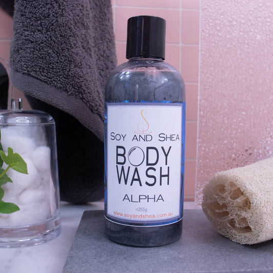 Bottle of "soy and shea body wash" on a bathroom counter, accompanied by a loofah, a towel, and a glass jar of cotton balls.