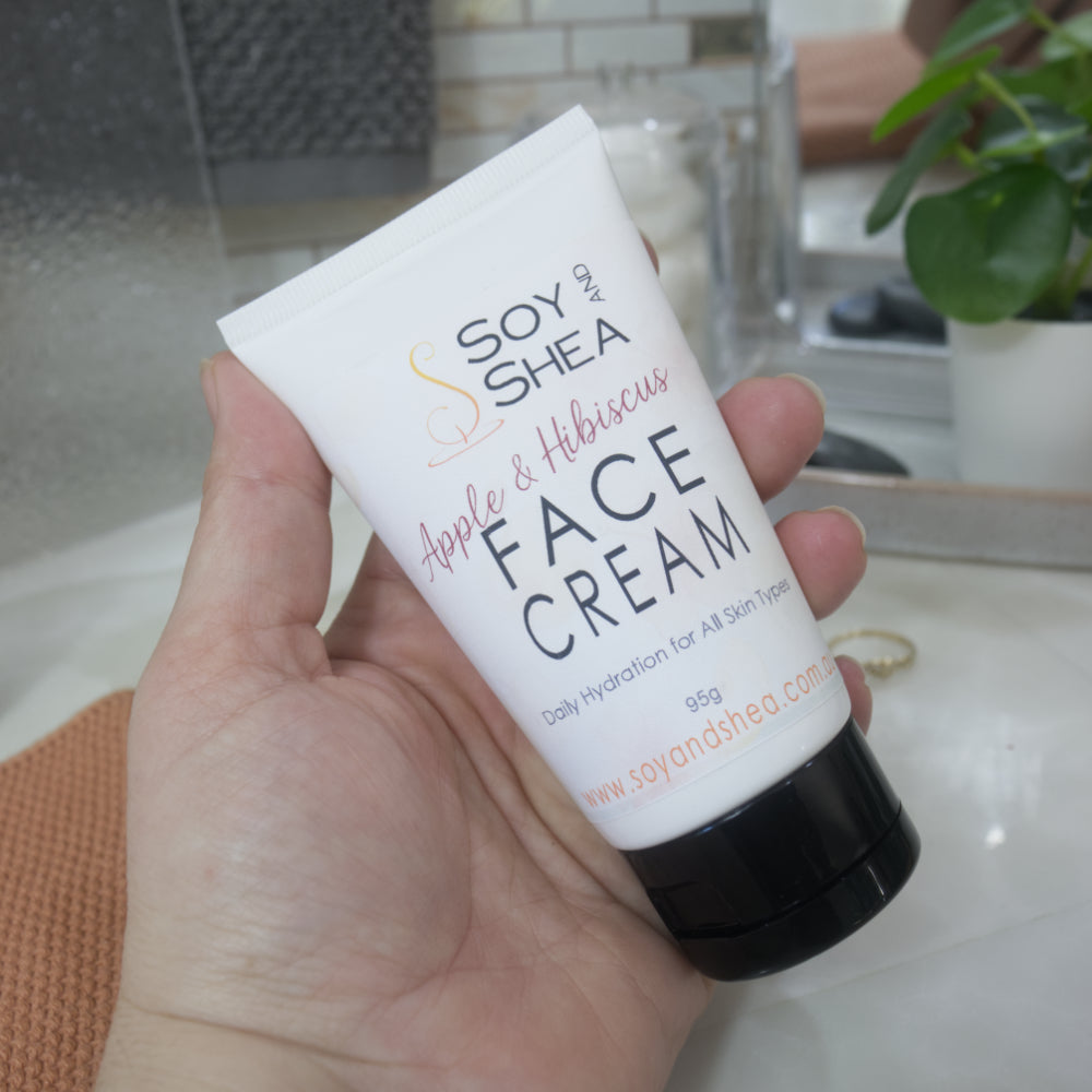 A hand holding a white squeeze tube of Soy and Shea Apple & Hibiscus Face Cream with a black cap, set against a softly blurred bathroom background.