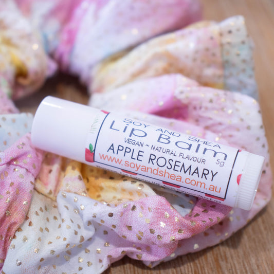 A white tube of lip balm sits on top of a scrunchie in pale rainbow colours.  The label reads LIP BALM APPLE ROSEMARY and has a white background with apple illustrations.