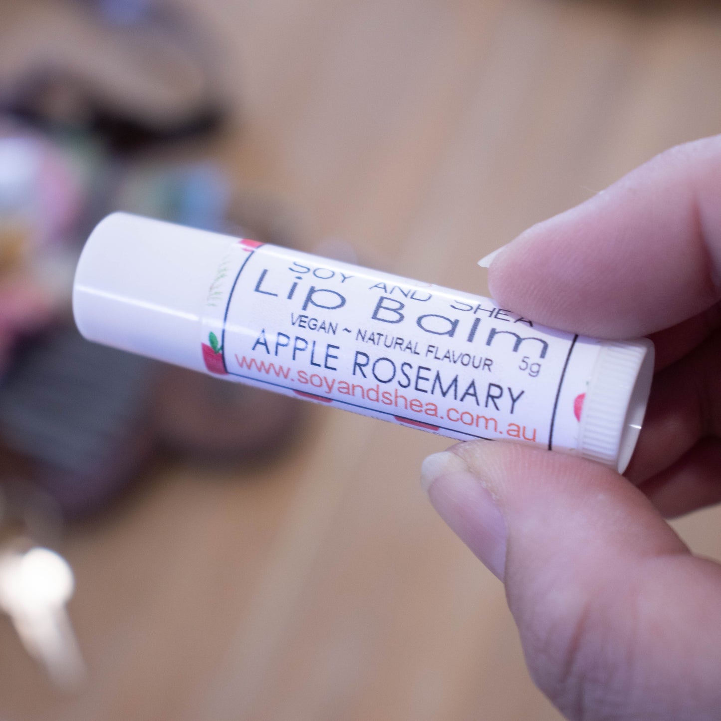 A hand holds a white tube labelled lip balm over a blurred image of an open handbag with contents spilling onto the table.  The lip balms features a while label with illustrations of red apples