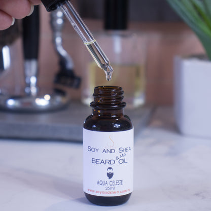 A small brown bottle labeled "Soy and Shea Midnight Masquerade Beard & Mo Oil," sits on the marble surface with shaving accessories on a blurred background.  A hand holds the dropper lid above the opening of bottle dripping the clear oil into the bottle