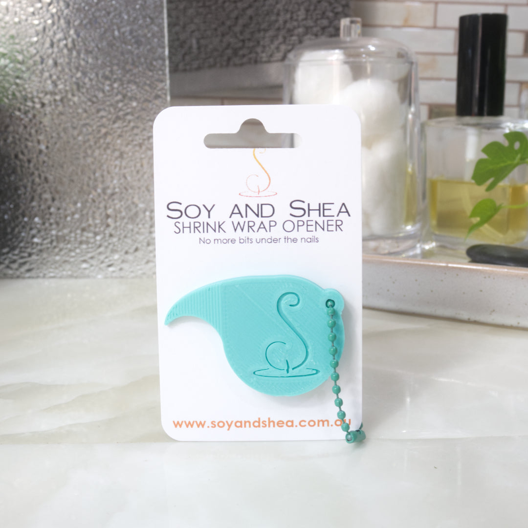 An Aqua Shrinkwrap opener with matching ball and chain keyring is attached to a white card with product details.  The card is standing upright on a bathroom countertop with a bottle of perfume, a jar of cotton balls and towel in the background