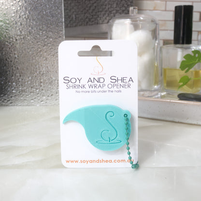 An Aqua Shrinkwrap opener with matching ball and chain keyring is attached to a white card with product details.  The card is standing upright on a bathroom countertop with a bottle of perfume, a jar of cotton balls and towel in the background