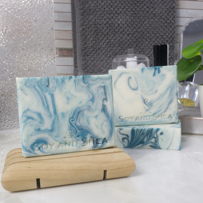 Three rectangular bars of soap are presented on a bathroom counter.  One soap is displayed standing upright on a wooden soap rack while to the back right, another soap lays flat revealing the soap top with a third standing upright on it.  The soaps all have a white base with varying swirls of teal throughout.  The soaps have a flat soap with swirling colour pattern