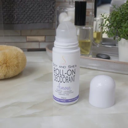 A white bottle of roll on deodorant stands on a bathroom countertop along with a body brush, bottle of perfume and a plant.  The domed lid has been removed and sits the side revealing a ball roller in the top of the bottle