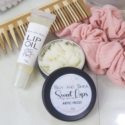 A small black tin with the lid removed revealing a white Lip Scrub lies on a marble tile surface next to a small, clear tube of white Lip Oil.   This rests againsts a wooden comb and pink scrunchie. 