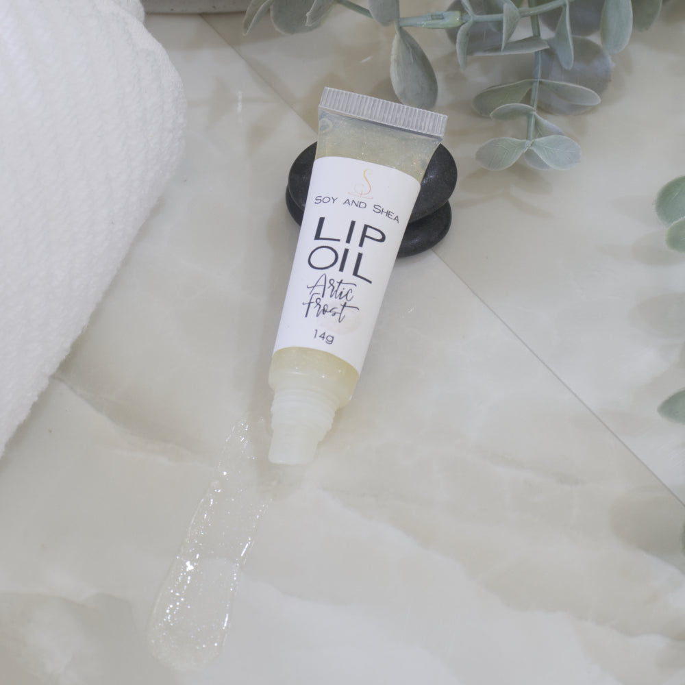 A tube of Lip Oil lies on a marble tile surface next to a white towel and some eucalyptus leaves.   The tube is clear with a white product label and filled with an off while Lip oil.  The cap is removed and a smear of Lip oil is on the bench showing the translucent effect it has