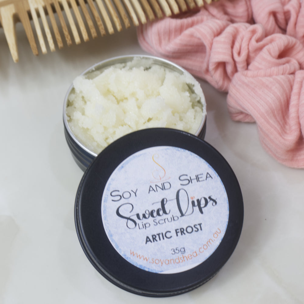 A black tin of Lip Scrub sits on a marble countertop next to a scrunchie and wooden comb.  The lid has been removed and rests on the side of the tin revealing a white scrub