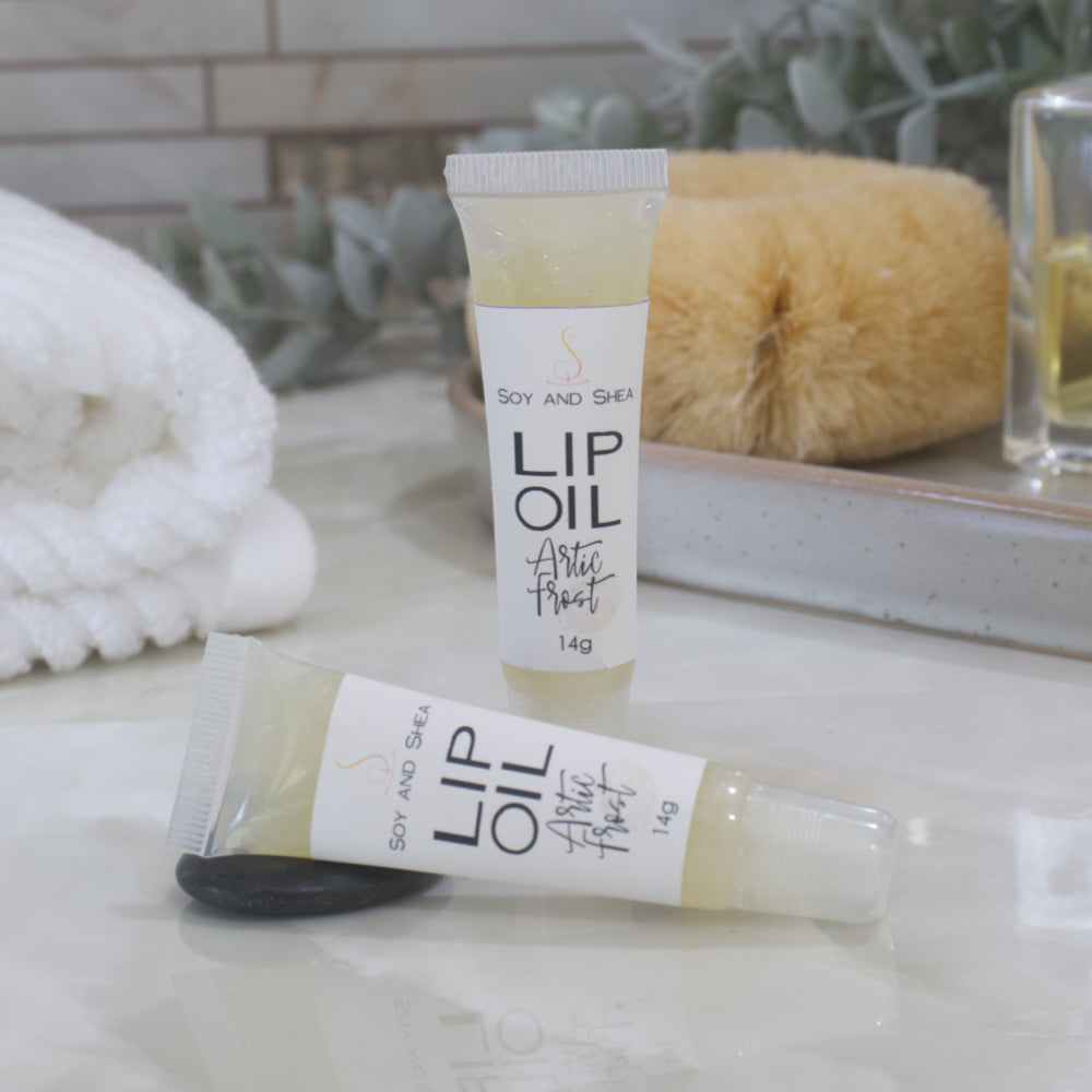 Two tubes of Lip Oil are presented on a bathroom countertop with a blurred background of towels, greenery and body brush.   One tube stands upright while the other lays flat in front of it.  The tubes are clear with a white product lable and are filled with an off white Lip oil which appears to have a slight shimmer.