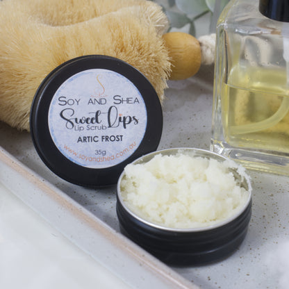 A round black tin sits on a stone tray along with a bottle of perfume and body brush.  The lid has been removed and is resting against the body brush revealing a white  sugar scrub 