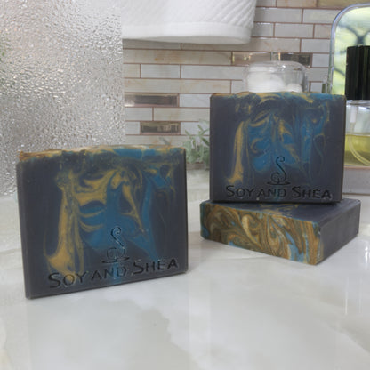 Three bars of Soap and Shea soap on a bathroom counter with a tiled wall in the background.  One soap lays flat on the surface showing a top that had dark gold and blue swirls.  A second soap stands upright on this soap with a third to the front-left.  These soaps are dark blue with bright blue and gold  swirls of colour dropped through.