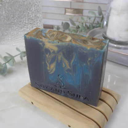 A dark blue soap bar with gold and bright blue drops of swirled colour stands on a wooden soap dish on a bathroom counter top.  